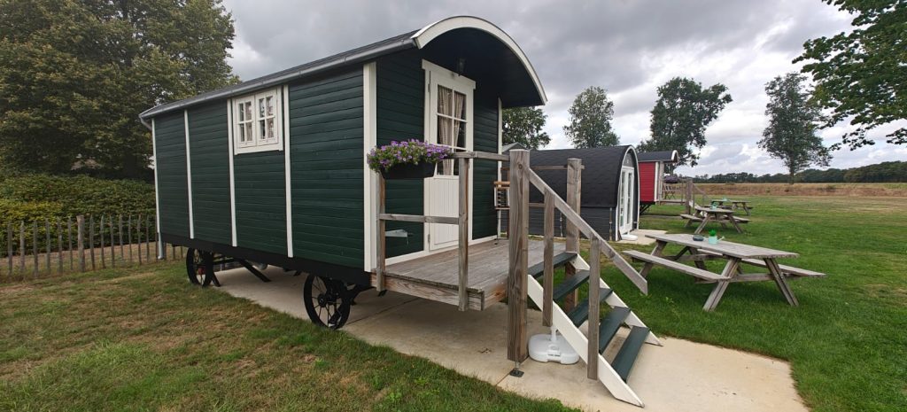Donkergroene pipowagen van opzij met houten wielen en daarachter twee camping pods met picknicktafels