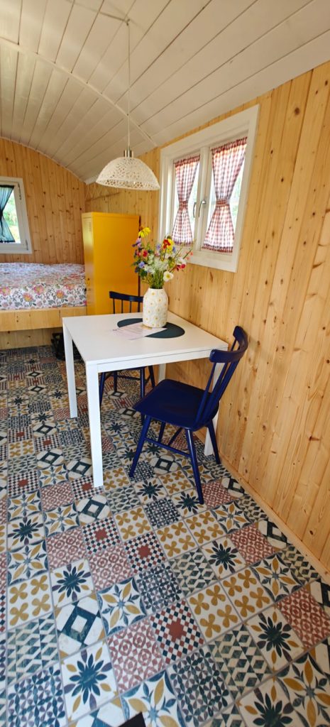 Interieur van de pipowagen met witte eettafel, blauwe stoelen, portugese tegelvloer en bed