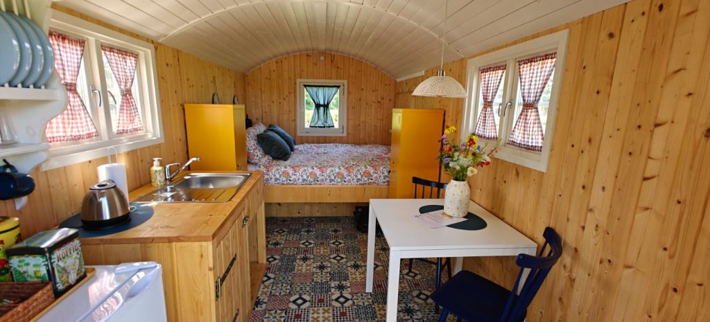 Interieur van de pipowagen met keukenblok, opgemaakt bed en eettafel onder een houten tongewelf